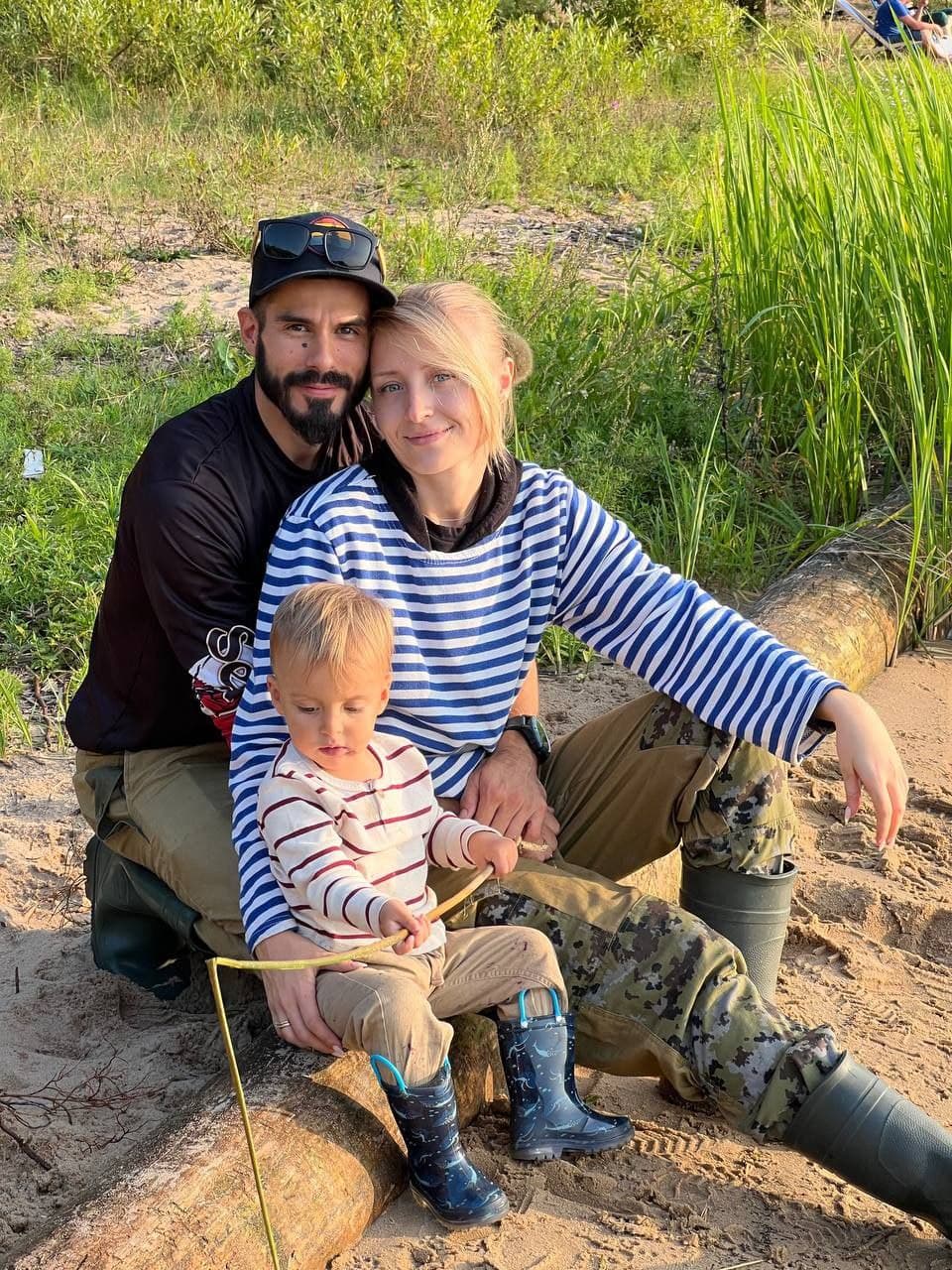 A family with a small child sits on a sandy riverbank near tall green grass.