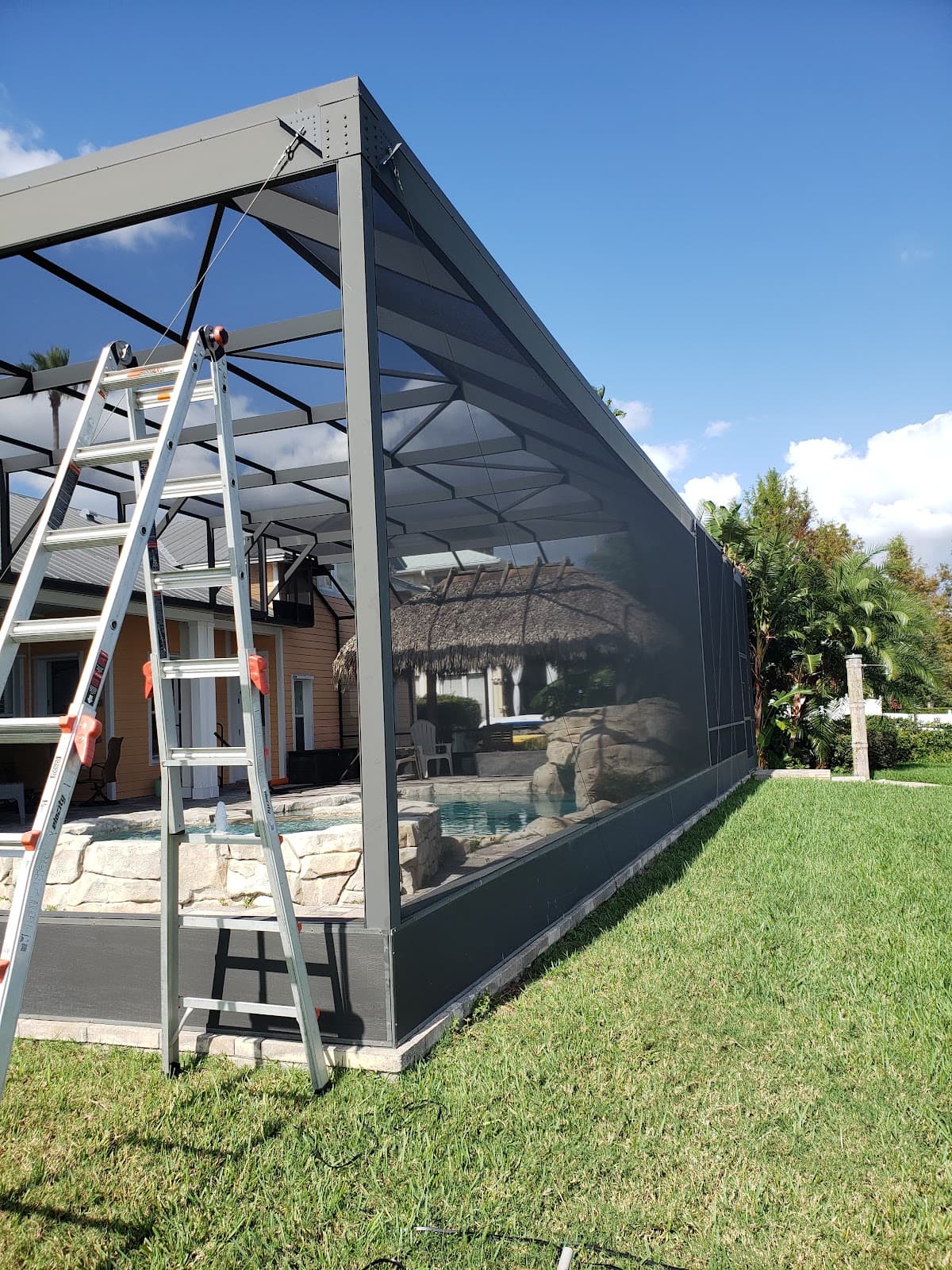 Silver ladder leaning against a grey screened pool enclosure featuring a thatched tiki hut inside.