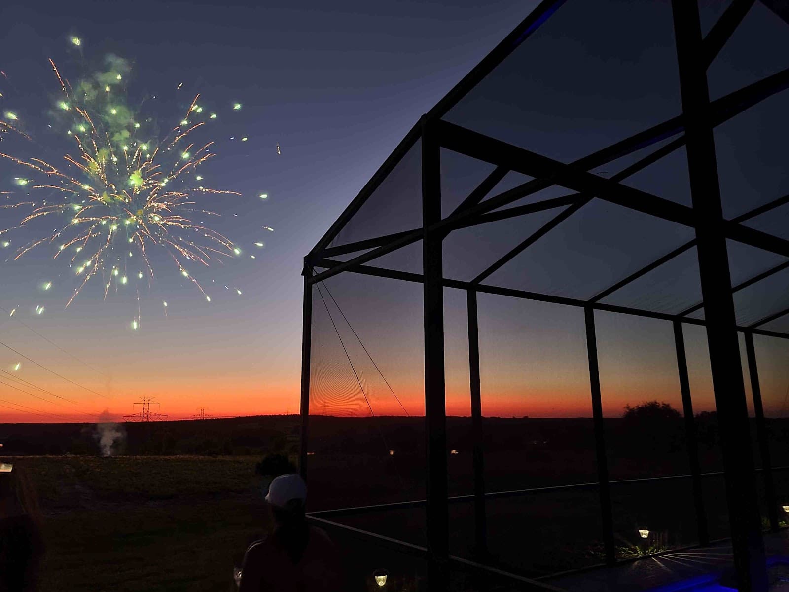 Green fireworks burst in a twilight sky over a sunset horizon next to a screened enclosure.