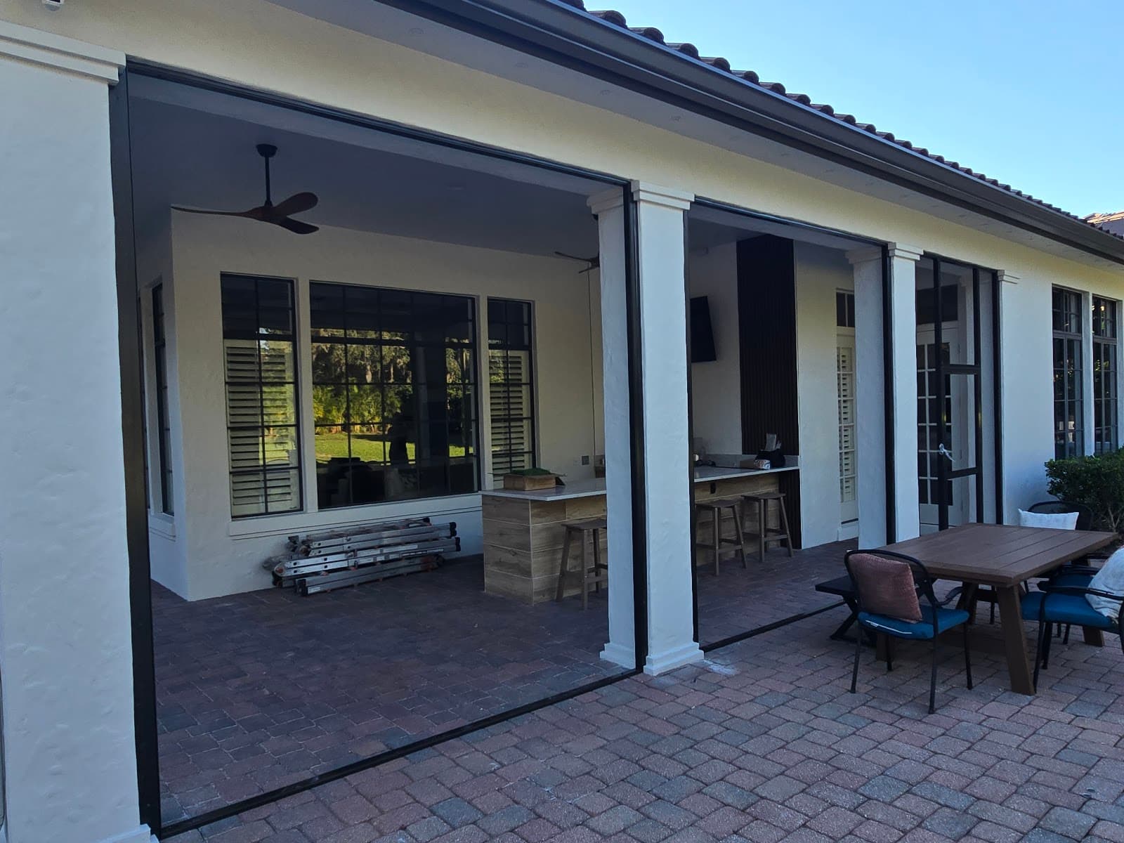 Modern white patio featuring retractable black screens, brick pavers, and an outdoor kitchen with stools.